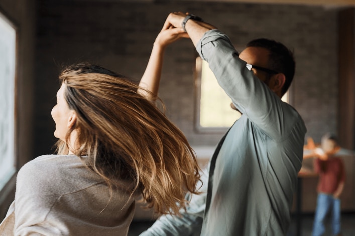 Deux personnes dansent dans une pièce avec fenêtres, autres en arrière-plan.