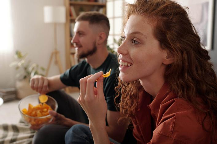 Seitenansicht einer lächelnden jungen Frau mit langen roten Locken, die knusprige Kartoffelchips isst und einen Film schaut