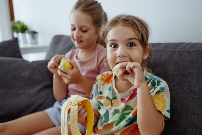 Zwei Mädchen essen Obst – eines schaut lächelnd mit einer Banane