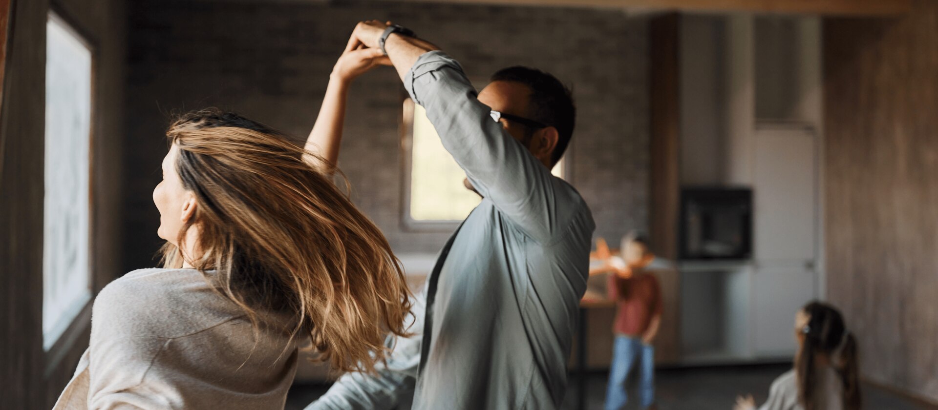 Deux personnes dansent dans une pièce avec fenêtres, autres en arrière-plan.