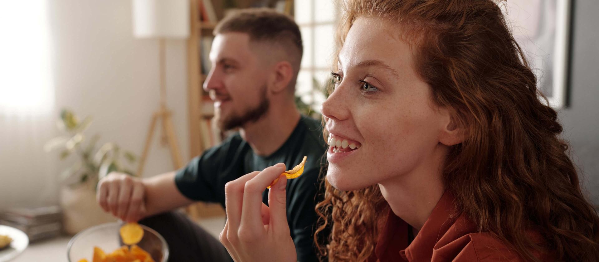Profil d'une jeune femme souriante avec de longs cheveux roux ondulés, dégustant des chips croustillantes en regardant un fil