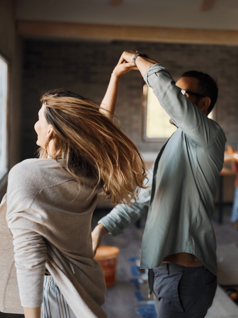 Zwei Personen tanzen in einem Raum mit Fenstern, weitere im Hintergrund.