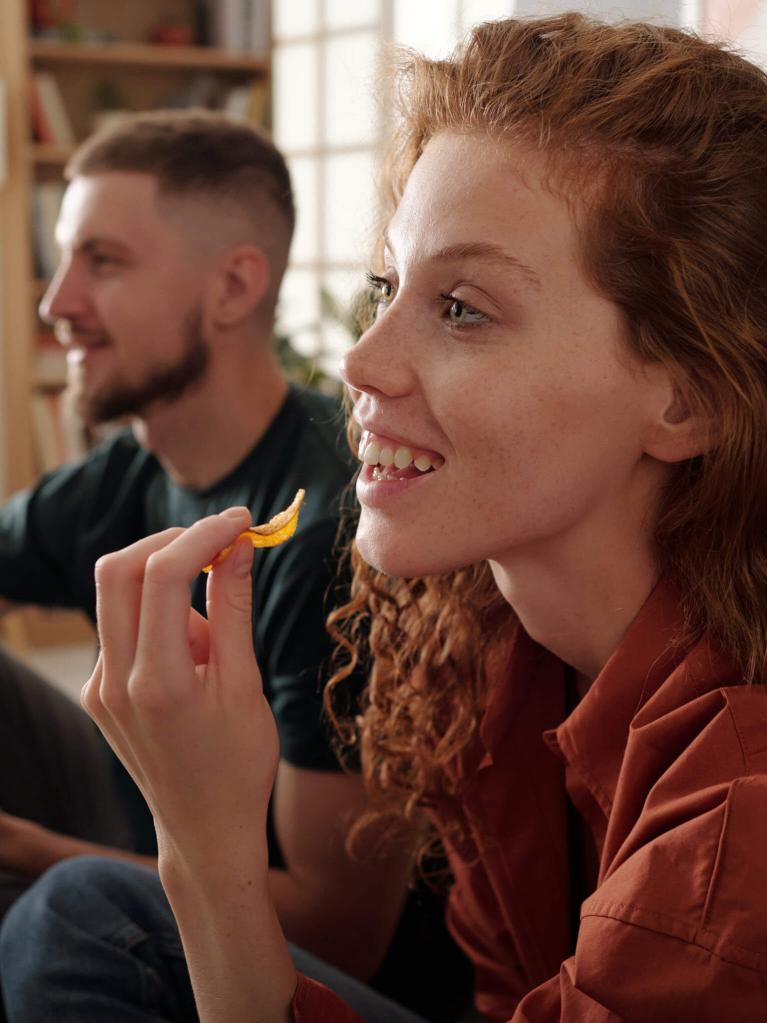 Profil d'une jeune femme souriante avec de longs cheveux roux ondulés, dégustant des chips croustillantes en regardant un fil