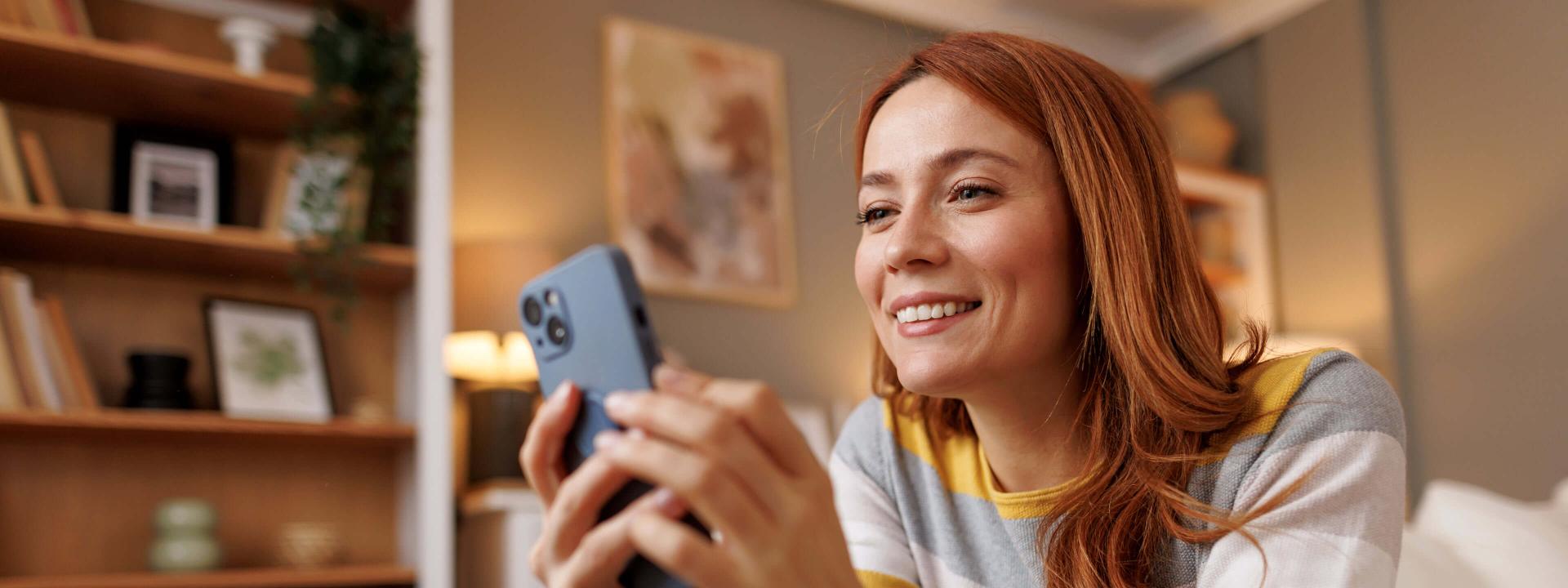 Jeune femme souriante allongée sur un canapé, utilisant son smartphone chez elle