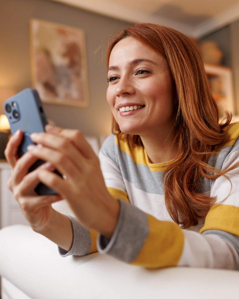 Jeune femme souriante allongée sur un canapé, utilisant son smartphone chez elle