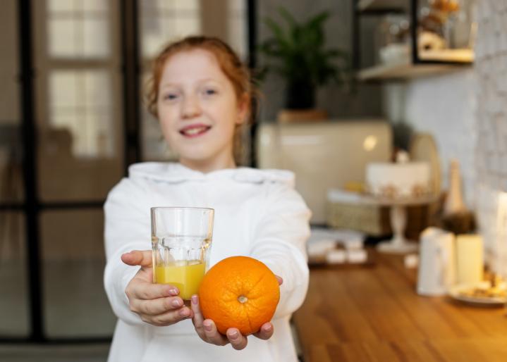 Junges rothaariges Mädchen in moderner Küche, spielt mit einer Orange und trinkt Orangensaft